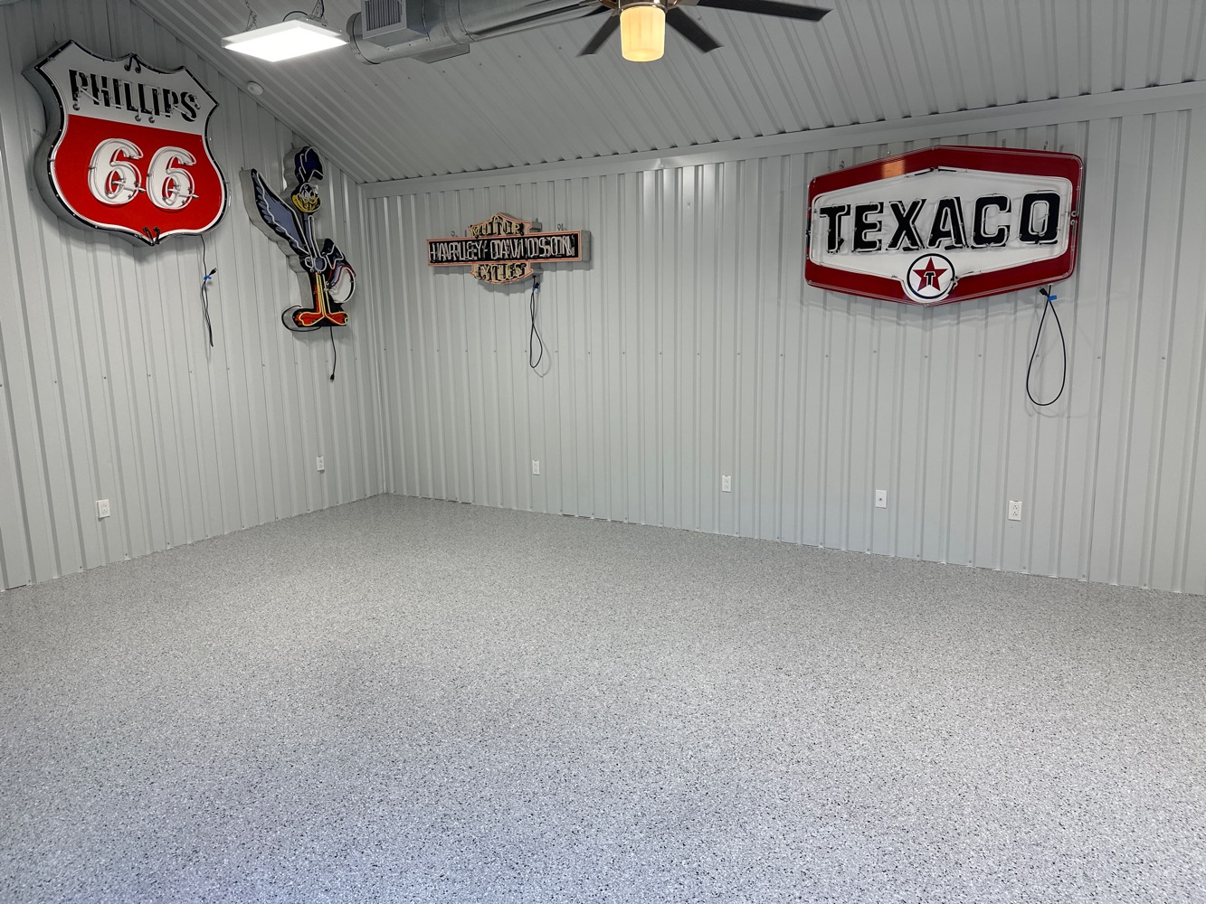 A clean, empty room with light gray speckled flooring and corrugated metal walls, showcasing vintage Phillips 66, Harley-Davidson, and Texaco signs, plus an image of a cartoon bird character—perfect for striking before and after photos.