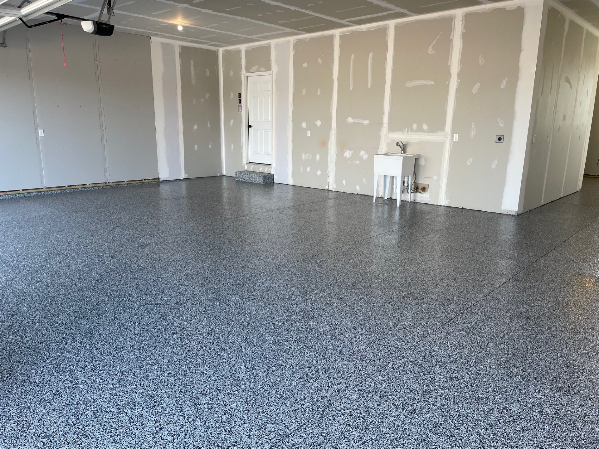 A clean, empty garage with gray speckled epoxy flooring, unfinished drywall, a white door with steps, and a small utility sink against the back wall—perfect for stunning before and after transformations. The space is well-lit and organized.