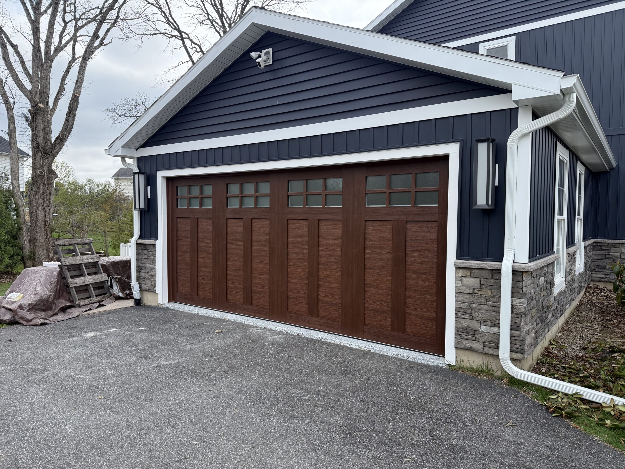 A two-car garage with a dark brown wooden door featuring windows at the top, attached to a blue house with stone accents and white trim. There is a covered pile of materials and wooden planks next to the garage.