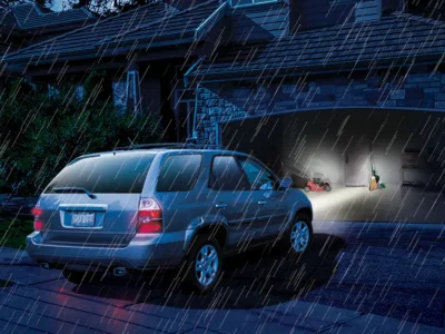 A silver SUV is stopped in front of an open garage on a rainy night. The garage door is illuminated with light, revealing scattered items and a person inside working or searching for something. Raindrops are visible in the light from the garage.
