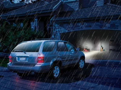 StormProofPower A silver SUV is stopped in front of an open garage on a rainy night. The garage door is illuminated with light, revealing scattered items and a person inside working or searching for something. Raindrops are visible in the light from the garage.