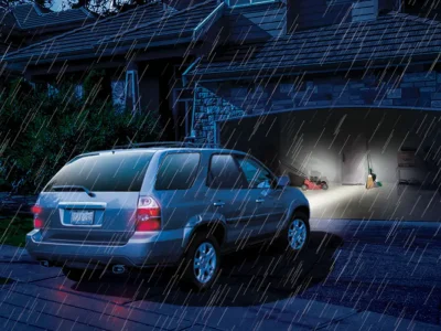 StormProofPower A silver SUV is stopped in front of an open garage on a rainy night. The garage door is illuminated with light, revealing scattered items and a person inside working or searching for something. Raindrops are visible in the light from the garage.